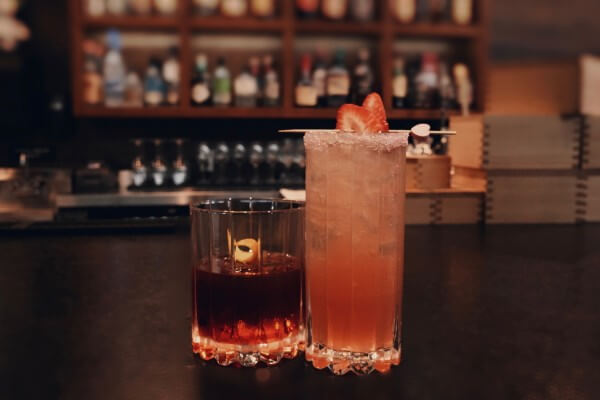 Two cocktails on the bar — an old fashioned and a tall strawberry-garnished highball